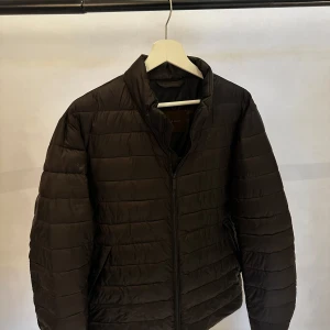 Svart dunjacka från Massimo Dutti - Snygg svart dunjacka från Massimo Dutti med quiltad design och hög krage. Jackan har dragkedja framtill, tryckknappar vid ärmsluten och två sidofickor. Perfekt för kyliga dagar och enkel att matcha med allt.