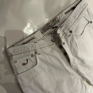 Vita jeans från Jacob Cohen, strl 34 - Snygga vita jeans från Jacob Cohen, modell Type 688. Byxorna har bruna kontrastsömmar, klassisk femficksdesign och detaljerade broderier. Tillverkade i bomull med Jacob Cohens ikoniska patch bak. Perfekta för dig som vill sticka ut med stil.
