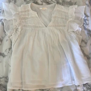 Vit blus med volangärmar Gina Tricot - Supersöt vit blus från Gina Tricot med volangiga ärmar och v-ringning. Blusen har broderade detaljer och är tillverkad i ett lätt, luftigt tyg som känns perfekt för varmare dagar. Använd ett fåtal gånger och är i storlek 146/152