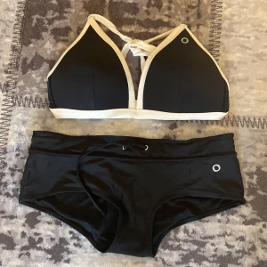 Svart bikini från Röhnisch med vita detaljer - Snygg svart bikini från Röhnisch med vita kanter och coola silverfärgade metalldetaljer. Överdelen (stl.L) har snörning i ryggen och sportig look, medan underdelen (stl M) har dragsko i midjan. Materialet är stretchigt och bekvämt, perfekt för strand eller poolhäng.