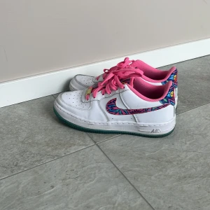 Nike Air Force 1 med färgglada detaljer - Unika Nike Air Force 1 sneakers i vitt skinn med färgglada, grafiska mönster på swoosh och häl. Skorna har rosa snören och foder, samt blå sula. 