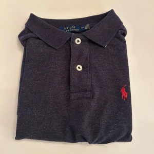 Mörkblå pikétröja från Ralph Lauren - Snygg mörkblå pikétröja från Polo Ralph Lauren med klassisk krage, två knappar och röd broderad logga på bröstet. Modellen är custom slim fit och passar perfekt till jeans eller shorts. Materialet är mjukt och skönt i bomull.