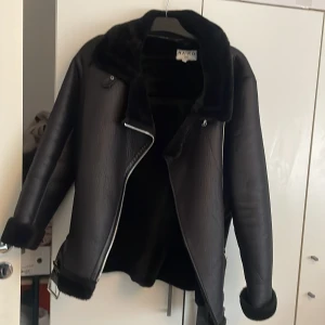 Svart pälsjacka från NA-KD med bälte - Snygg svart pälsjacka från NA-KD med mjuk fuskpäls på insidan och kragen. Jackan har dragkedja, bälte i midjan och detaljer vid ärmsluten. Perfekt för kalla dagar när du vill vara både varm och stilren.