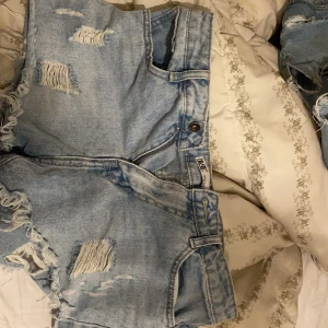 Ljusblåa jeansshorts med slitningar - Säljer ett par ljusblåa jeansshorts från INSF med snygga slitningar och fransiga kanter. Shortsen har klassisk femficksdesign och knappstängning framtill. Perfekta för varma dagar när du vill ha en avslappnad och trendig look.