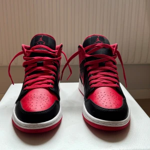 Nike Air Jordan 1 Mid Svart/Röd - Säljer ett par Nike Air Jordan 1 Mid i svart och rött skinn med klassisk vit sula och röda snören. Ikonisk siluett med perforerad tå, röda detaljer och Jordan Wings-logga på sidan. Perfekta för dig som vill sticka ut med en tidlös sneaker.