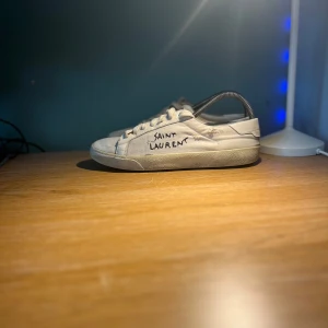 Saint Laurent skor - Saint Laurent sneakers i vit canvas med handskriven logga på sidan. Klassisk låg modell med snörning och rund tå. Snygg minimalistisk design och slitna detaljer för en cool vintagekänsla. Perfekta för dig som gillar streetstyle och designer vibes.