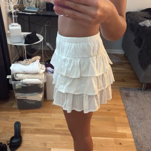 Vit volangkjol med resårmidja - Supersöt vit kortkjol med flera lager volanger och bred resår i midjan. Kjolen är luftig och har en flowy känsla, perfekt för dig som gillar en lekfull och trendig stil. .