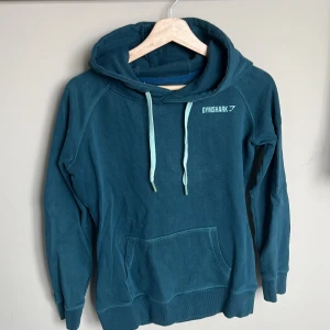 Blå hoodie från Gymshark med huva - Mysig blå hoodie från Gymshark med huva och dragsnören i ljusare blå nyans. Tröjan har en stor magficka och långa ärmar med muddar. Tröjan är något nopprig och gummit är något missfärgat längts ut på snörena. Det står ingen storlek i tröjan men den passar dig som har storlek S.