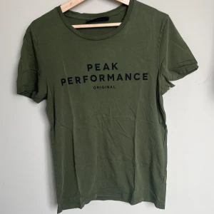 Grön Peak Performance t-shirt i bomull - Snygg grön t-shirt från Peak Performance med svart tryck på bröstet. Klassisk rund halsringning och korta ärmar. Tillverkad i mjuk bomull. Förekommer två små solblekta fläckar på framsidan, men som bara syns om man kollar noga.