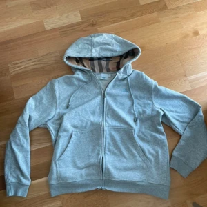 Grå hoodie från Burberry med dragkedja - Säljer en grå hoodie från Burberry i bomull med klassiskt randigt foder i huvan. Tröjan har dragkedja, snörning i huvan och broderad Burberry-logga på bröstet. Perfekt för dig som gillar stilrena och exklusiva plagg.