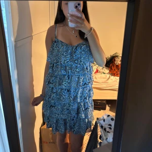 Blå mönstrad volangklänning - Söt blå kortklänning med paisleymönster och volanger. Klänningen har tunna axelband och en luftig passform, perfekt för varma dagar. Flera lager volanger ger en lekfull känsla och den är superbekväm att bära. stl s/m skulle jag säga! men står ingen lapp eller så
