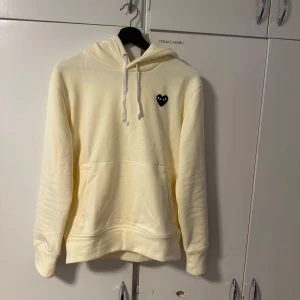 Gul hoodie från Comme des Garçons Play - Snygg ljusgul hoodie från Comme des Garçons Play med klassiskt svart hjärta-logga på bröstet. Tröjan har huva med vita snören, känguruficka och långa ärmar. Perfekt för dig som gillar stilrena och trendiga plagg med en twist.