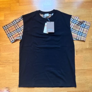 Burberry t-shirt med rutiga ärmar - Svart t-shirt från Burberry med klassiska rutiga ärmar i beige, svart, vitt och rött. T-shirten har rund halsringning och är tillverkad i mjuk bomull. Perfekt statement-plagg med ikonisk design och snygg kontrast mellan ärmar och kropp.