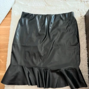 Svart volangkjol i skinnimitation - Snygg svart kjol i skinnimitation med volang nertill. Kjolen är kort och har en tight passform som framhäver figuren. Perfekt för dig som vill ha en edgy och trendig look.