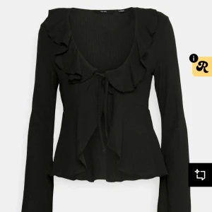 Svart kofta med volang och knyt - Supersnygg svart kofta från Vila med volangdetaljer längs ringningen och knyt framtill. Långärmad och lätt croppad passform som ger en trendig siluett. Perfekt att styla med jeans eller kjol för en cool look.