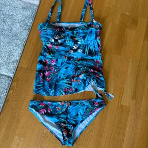 Blommig blå bikini med snörning - Snygg bikini i blått med tropiskt blommönster i rosa, vitt och grönt. Överdelen har breda axelband och en längre modell med snörning i sidan för extra detalj. Underdelen är klassisk med samma färgglada blommiga print. Perfekt för strandhäng och poolparty!
