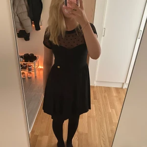 Svart kortklänning med prickig mesh - Supersöt svart kortklänning med transparent mesh upptill och prickigt mönster. Klänningen har rund hals, korta ärmar och markerad midja. Kjolen är lätt utställd och en liten guldfärgad knappdetalj sitter i midjan. Perfekt för dig som gillar en lekfull men stilren look.