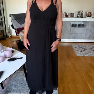 Svart långklänning med smala axelband - Snygg svart långklänning med smala axelband och v-ringad omlott framtill. Klänningen har markerad midja och ett lätt, flytande tyg som ger en elegant siluett. Perfekt för dig som gillar stilrena och klassiska plagg med en modern touch.