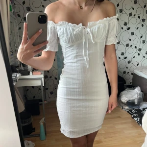 Vit offshoulder kortklänning BikBok - Supersöt vit kortklänning från BikBok med offshoulder och smockad struktur. Klänningen har korta puffärmar, söt knytning framtill och en figurnära passform. Perfekt för sommarens alla tillfällen! Funkar även jättebra som klänning till studenten eller examen.