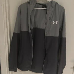 Grå vindjacka från Under Armour - Snygg tvåfärgad vindjacka från Under Armour i ljusgrått och mörkgrått. Jackan har huva, dragkedja framtill, mesh-detaljer på sidorna och en bröstficka med dragkedja. Perfekt för dig som gillar sportig stil och vill ha något lätt och funktionellt.