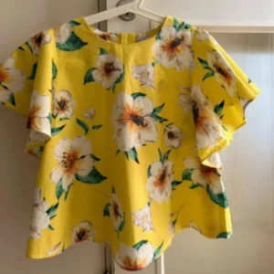 Ny Gul blommig blus med vida ärmar - Superfin gul blus med stora vita blommor och gröna blad. Blusen har korta, vida ärmar och knappar längs ryggen. Perfekt för dig som vill sticka ut med färg och mönster. Luftig och skön modell som passar till jeans eller kjol. 