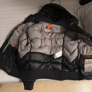 Svart dunjacka Parajumpers med huva - Säljer en svart jacka frän Parajumpers med huva och breda quiltade paneler. Jackan har grått innerfoder, ribbade muddar och klassisk logga på ärmen. Den har två hål på ärmarna som ni ser. Men vill bara bli av med jackan
