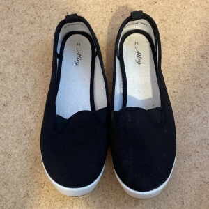 Svarta ballerinaskor från Alley - Svarta ballerinaskor från Alley i textil med vit platt sula. Skorna har en enkel och stilren design med rund tå och låg profil, perfekta för en avslappnad look. Lätta att ta på och av tack vare den flexibla öppningen.