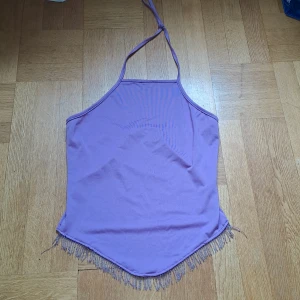Lila halterneck topp med fransar  - Säljer en lila halterneck linnetopp från Lucy Paris. Toppen har smala axelband som knyts i nacken och en spetsig nederkant med fransdetaljer. Materialet är mjukt och stretchigt. Uppskattad storlek s. 