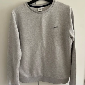 Grå sweatshirt från BOSS - Snygg grå sweatshirt från BOSS med diskret logga på bröstet. Tröjan har rund halsringning, långa ärmar och ribbade muddar. Perfekt för en clean och stilren look. Materialet är mjukt och skönt, troligtvis bomullsmix. Tröja är i nyskick!