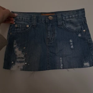 Blå jeanskjol med nitar - Säljer en kort blå jeanskjol från Lizabeta jeans med slitna detaljer och coola nitar både fram och på bakfickorna. Kjolen har klassiska fickor och bälteshällor, perfekt för en elegant look. (Säljer den för att den var för liten, den är helt nytvättad och oanvänd) 🩵