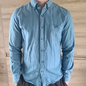 Blå skjorta Dressmann - Snygg blå skjorta från Dressmann i slim fit-modell. Skjortan har klassisk krage, bröstficka och knäppning framtill samt vid ärmslut. Tillverkad i 100% mjuk bomull, perfekt för en clean och stilren look. Inga defekter, kan behöva strykas bara.