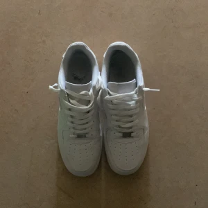Nike Air Force 1 - Klassiska Nike Air Force 1 i helvitt skinn med perforerad tå, rund tå och platt sula. Snygg low-top modell med snörning och diskret Nike-logga på sidan. Perfekta för dig som gillar en clean och tidlös look.