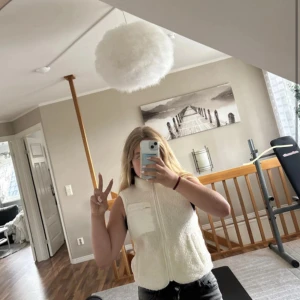 Vit fleeceväst från Lindex Kids - Mysig vit fleeceväst från Lindex Kids med hög krage och dragkedja framtill. Västen har en slät bröstficka med dragkedja och två sidofickor. Perfekt för lager-på-lager och enkel att matcha med andra plagg.134/140 i storlek