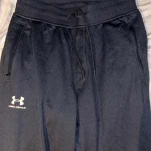 Svarta tränings från Under Armour - Svarta mjukisbyxor från Under Armour med dragsko i midjan och dragkedjefickor på sidorna. Byxorna har en sportig look med logga på vänster ben och är tillverkade i ett mjukt, syntetiskt material som passar perfekt för träning eller chill.