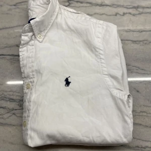 Vit skjorta från Ralph Lauren - Klassisk vit skjorta från Ralph Lauren med broderad marinblå logga på bröstet. Skjortan har knappar framtill och krage, samt är tillverkad i bomull. Perfekt för en clean och stilren look.