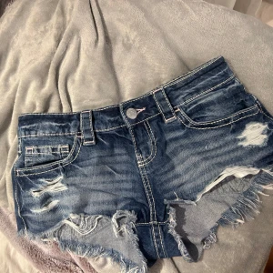 Blå slitna jeansshorts med fransar - Snygga blå jeansshorts med slitna detaljer och fransig kant. Shortsen har klassiska fickor fram och bak, markerade sömmar och låg midja. Perfekta för sommardagar när du vill ha en avslappnad och trendig look. Går inte under priset då dom är helt nya❤️