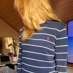 Randig blå stickad tröja från Ralph Lauren - Mörkblå stickad tröja från Ralph Lauren med vita horisontella ränder och rosa broderad logga på bröstet. Tröjan har rund halsringning, långa ärmar och ribbade muddar. Perfekt för dig som gillar klassisk och stilren look.