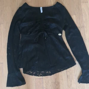 Svart spetsblus - Supersnygg svart blus från Chiara Forthi med spetsdetaljer och lång ärm. Blusen har v-ringning och snörning framtill som ger en cool look. Perfekt för dig som gillar feminina detaljer och vill sticka ut lite extra.