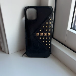 Svart mobilplånbok Ideal of Sweden - Snygg svart mobilplånbok från Ideal of Sweden i skinnimitation med guldiga nitar i coolt mönster på framsidan. Plånboksmodellen har plats för mobilen och ger en edgy vibe med sina detaljer. Perfekt för dig som gillar accessoarer med attityd.