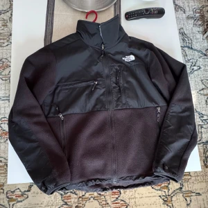 Svart fleecejacka från The North Face - Svart fleecejacka från The North Face med hög krage och dragkedja framtill. Jackan har flera praktiska fickor med dragkedja och en stilren design med förstärkta partier över bröst och axlar. Perfekt för kyliga dagar och outdoor vibes.