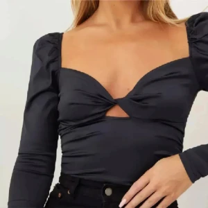 Svart omlottopp med puffärm - Snygg svart topp med omlottkänsla och twistad detalj framtill. Djup urringning, lång ärm med puff vid axlarna och liten cut-out under bysten. Sitter tight och har en glansig finish som ger en cool look. Tryck ej köp nu