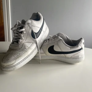 Nike Air Force 1 vita sneakers - Klassiska Nike Air Force 1 sneakers i vitt skinn med svart swoosh på sidorna. Lågt skaft, perforerad tå och platt sula. Perfekta för dig som gillar en clean och tidlös streetstil. Skorna har snörning och är riktigt ikoniska i sneaker-världen. Kan hämtas upp i Huddinge annars står man för frakten själv