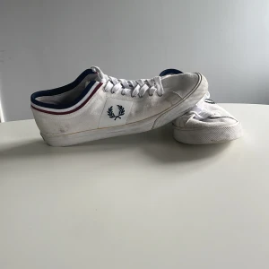 Vita sneakers från Fred Perry - Klassiska vita sneakers från Fred Perry i canvas med snörning. Skorna har marinblå och röda detaljer runt kragen samt Fred Perry-loggan på sidan och hälen. Platt sula och rund tå ger en clean och sportig look. Går att hämta upp i Huddinge annars står för frakten själv.