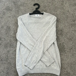 Ljusgrå tröja från Bläck, storlek M - En stilren ljusgrå tröja från Bläck i storlek M. Tröjan har rund halsringning, långa ärmar och ribbade muddar vid ärmslut och nederkant. Perfekt för lager-på-lager och enkel att matcha med jeans eller chinos.