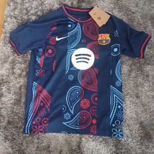 FC Barcelona Nike Dri-FIT fotbollströja - FC Barcelona matchtröja från Nike med Dri-FIT-material. Tröjan är marinblå med röda och ljusblå paisleymönster, klubbmärke på bröstet och Spotify-logga. Rund hals med röd och blå kant, kortärmad och tillverkad i funktionsmaterial.