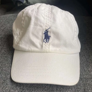 Ralph Lauren Keps Vit - Vit keps från Polo Ralph Lauren med klassisk blå broderad logga framtill och justerbart spänne bak. Kepsen är tillverkad i 100% bomull och har en stilren, tidlös design som passar till det mesta.