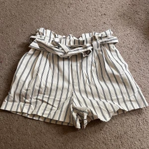 Randiga högmidjade shorts - Snygga vita shorts med svarta ränder och papperspåse-midja. De har ett matchande tygbälte och är perfekta för varma dagar. Modellen är lös och bekväm med trendig look.