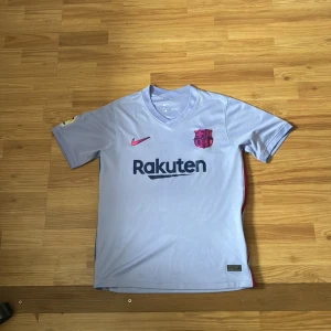 Barcelona bortatröja Nike Dri-FIT - Snygg Barcelona bortatröja från Nike med Dri-FIT-material. Tröjan är ljuslila med korta ärmar, V-ringning och tryckta loggor från Rakuten och UNICEF. FCB-emblemet i metallicrosa och Nike-logga på bröstet. Perfekt för dig som älskar fotboll och Barca. 