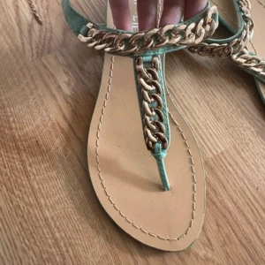 Turkos sandaler med guldkedja - Snygga sandaler i turkos med bred guldfärgad kedjedetalj över foten och runt vristen. Platt sula och öppen tå, perfekt för varma dagar. Remmarna är i syntetmaterial och sulan är beige. Kedjan ger en cool och trendig look