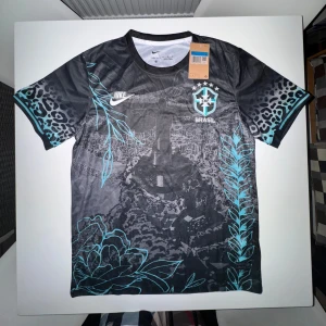 Nike x Brazil fotbollströja - Snygg svart Brasilien fotbollströja från Nike i STORLEK M! med unikt tryck av Kristusstatyn, blågröna blommor och blad samt leopardmönster på ärmarna. Tröjan har korta ärmar, rund hals och är tillverkad i lätt Dri-FIT material som andas. Perfekt för dig som vill sticka ut på planen.